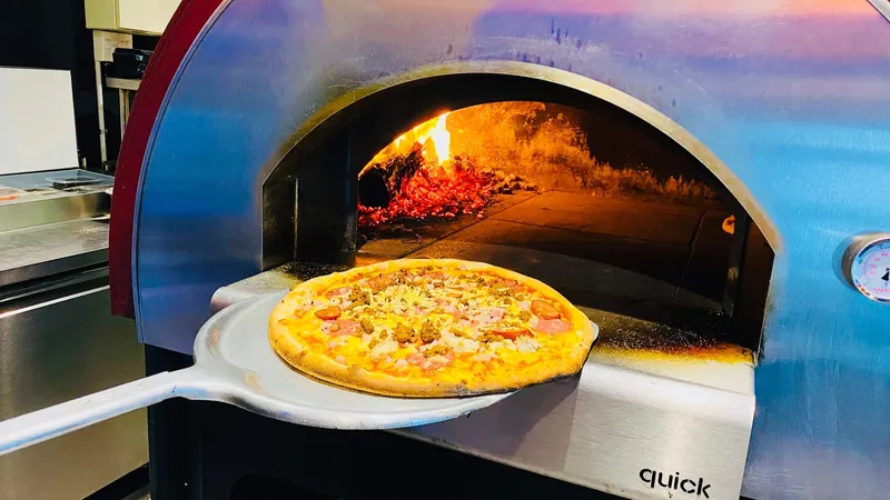 A pizza on a peel in front of Scoozi's lit wood-fired oven.