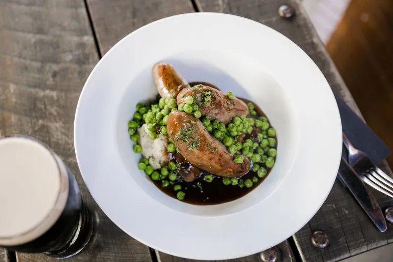 Bangers and mash with peas and onion gravy, photographed on a rustic table beside a dark beer.
