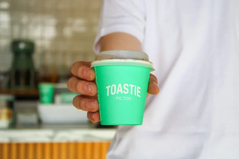 A hot drink in a bright green Toastie Picton branded takeaway cup held over the shop counter.